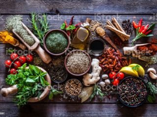 Top view of a rustic wood kitchen table filled with a large group of multi colored spices and herbs in bowls, wooden serving scoops or placed directly on the table. Spices and herb included are clove, turmeric, bay leaf, cinnamon, olive oil, curry powder, ginger, nutmeg, peppercorns, cinnamon, salt, chili pepper, basil, parsley, lemon, rosemary, garlic, onion and saffron. Low key DSRL studio photo taken with Canon EOS 5D Mk II and Canon EF 100mm f/2.8L Macro IS USM